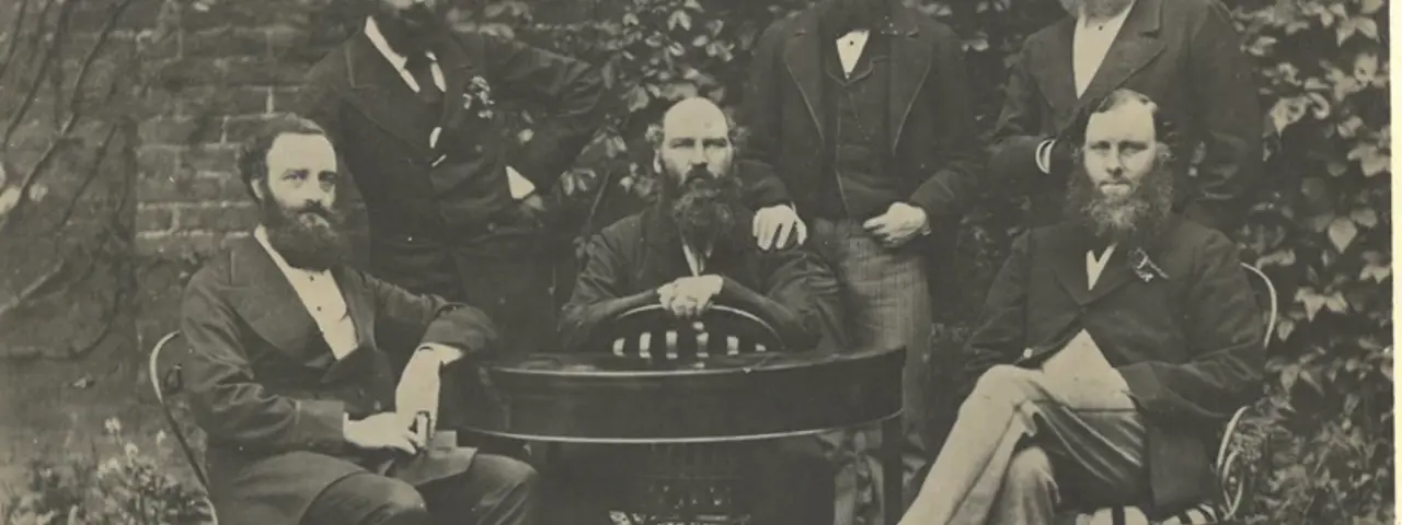 Black and white photo of six bearded men gathered around a table dressed in formal suits from the 1800s, threee seated and three standing